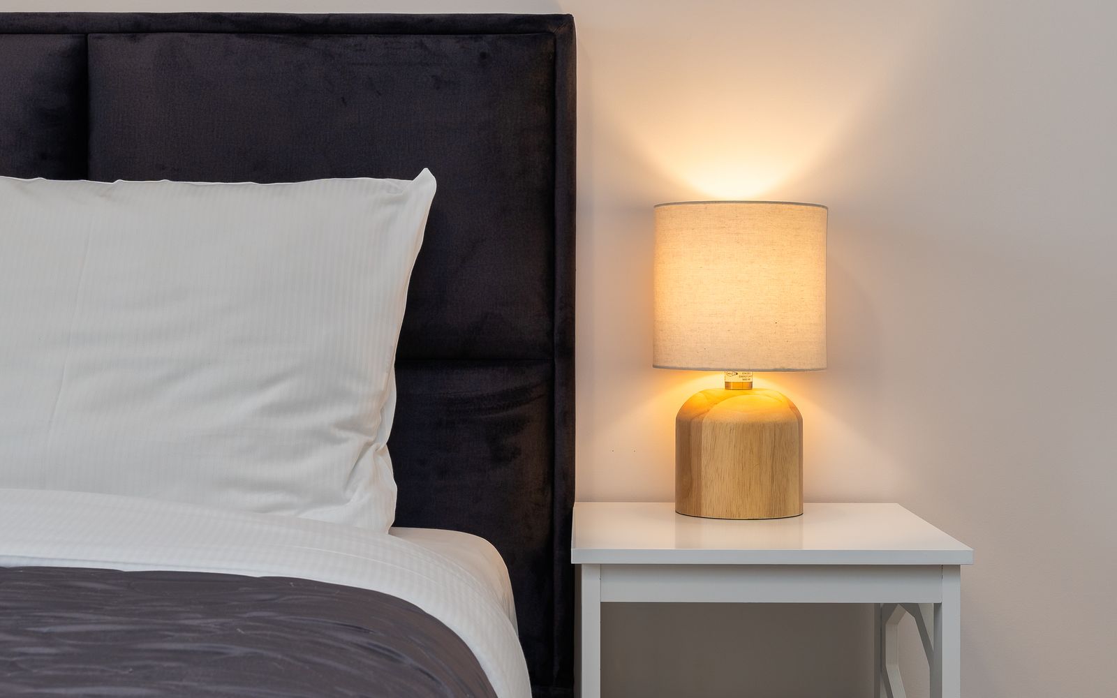 Lampada accesa su un comodino bianco accanto a un letto con cuscini bianchi, in una stanza con luce calda