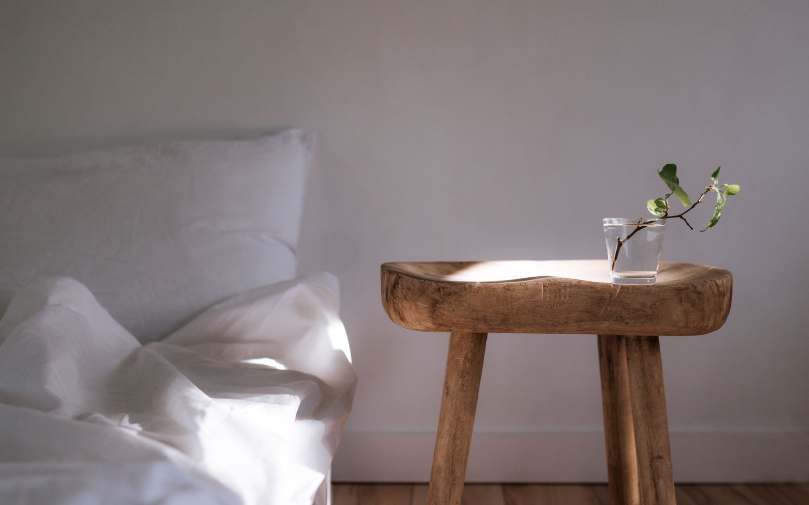Un comodino di legno con un bicchiere d'acqua e un rametto verde, accanto a un letto con lenzuola bianche, nella luce del mattino
