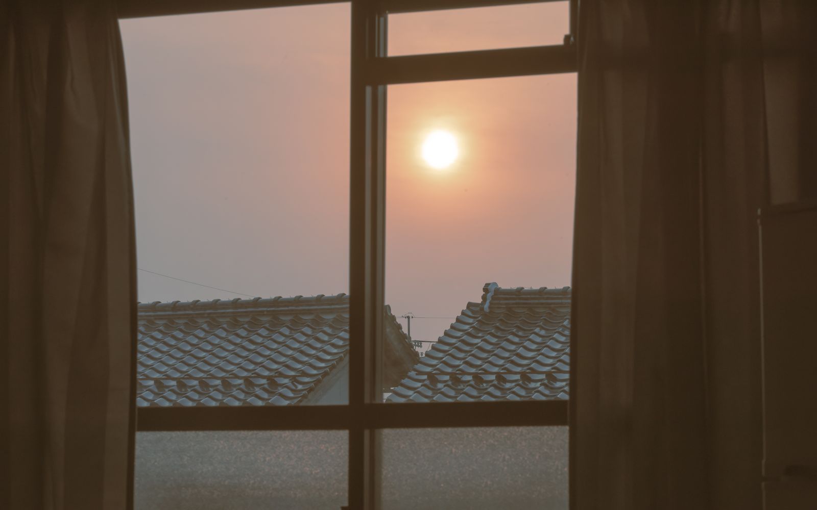 Una finestra con le tende scostate: il sole sorge sopra i tetti e il cielo è rosato